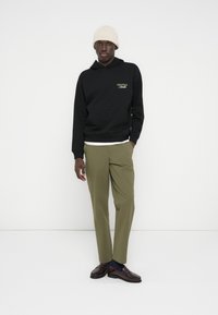 Άντρας που φοράει μπεζ σκουφάκι, μαύρο φούτερ με το κείμενο «Originals Studio», ελαιδί παντελόνι και σκουρό καφέ μοκασίνια, μπροστά σε λευκό φόντο.