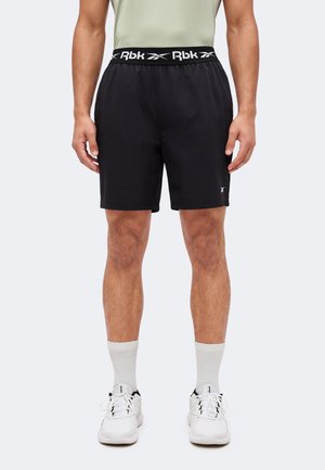 Mann trägt schwarze Reebok-Shorts mit Logo am Bund und am linken Bein, weiße Socken und weiße Sportschuhe, steht frontal.