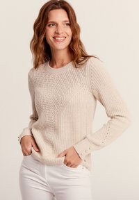 Pull en tricot beige avec un motif en losanges texturé, col rond et accents de boutons sur les poignets. Porté sur un pantalon blanc.