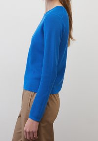 Donna che indossa un maglione lungo a maniche lunghe di un blu brillante e pantaloni marrone chiaro, in piedi di lato contro uno sfondo luminoso e semplice.