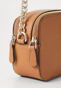 Tasche mit brauner Struktur, ausgestattet mit einer goldenen Kettenriemen, zwei Reißverschlüssen und schwarzem Piping. Kleine seitliche Akzente mit flachen Laschen für ein einfaches Öffnen.