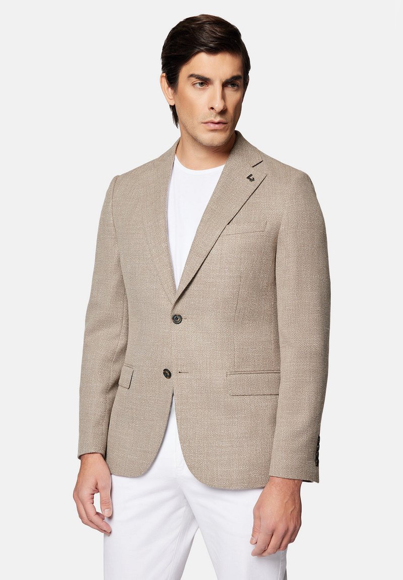 Beżowa marynarka w jodełkę z dwoma guzikami, klapami i dwoma przednimi kieszeniami. Nosi się ją na białej koszuli, w zestawie z białymi spodniami.