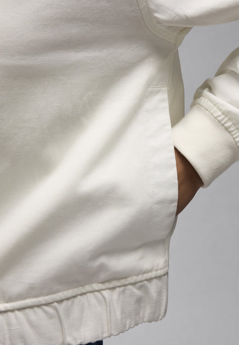 Veste en coton blanc avec un poignet côtelé et une poche latérale, au toucher lisse et avec des détails de couture sur l'ourlet.