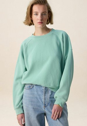 Femme portant un sweat-shirt oversize vert menthe et un jean bleu clair, debout devant un fond beige uni.