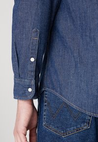 Denim overhemd met een puntige manchetknoop en witte knopen, lichtblauwe kleur, en contrasterende gele stiksels; vastgemaakt aan donkerblauwe jeans met gebogen zakken.