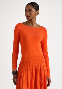 Moteris dėvi ryškios oranžinės spalvos ilgomis rankovėmis suknelę su klostuota suknele ir ant kairės riešo segi juodai auksinę apyrankę.