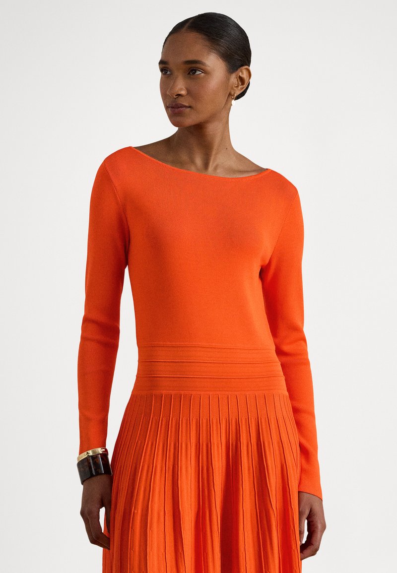 Moteris dėvi ryškios oranžinės spalvos ilgomis rankovėmis suknelę su klostuota suknele ir ant kairės riešo segi juodai auksinę apyrankę.