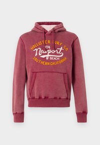 Sudadera burdeos con bolsillo delantero, capucha con cordón y gráfico en amarillo y blanco que dice "Hollister Surf Co Newport Beach 1974". Textura suave.