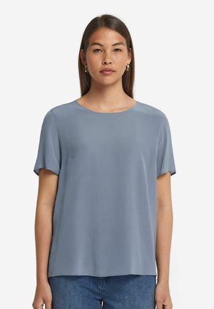 Helle blaue Bluse aus glattem Stoff, mit kurzen Ärmeln und Rundhalsausschnitt. Kombiniert mit blauen Denim-Jeans.