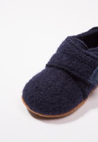 Granatowe filcowe kapcie z okrągłym noskiem, z rzepem i teksturowaną gumową podeszwą dla lepszej przyczepności. Miękki materiał zapewnia wygodę.