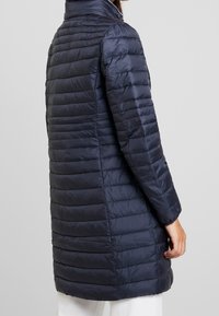 Marineblå pufferjakke med kviltet design, lynlåslukning, høj krave og lange ærmer. Glat, blankt stof med et let udseende.