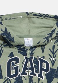 Zöld kisgyermek kapucnis pulóver tengerészkék leveles mintával és nagy tengerészkék "GAP" betűkkel az elején, cipzáros záródással, valamint belső címke látható.