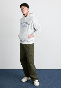 Siv hoodie z modrim napisom "New Balance Athletics", v kombinaciji z ohlapnimi olivno zelenimi vojaškimi hlačami in belimi supergami, stoji na modrem preprogi.