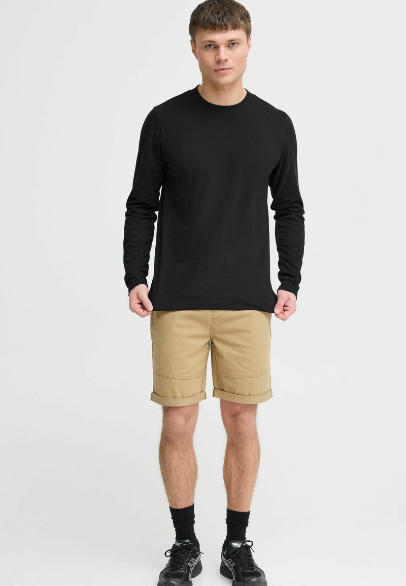 Zwarte long sleeve shirt, khaki shorts met opgerolde mouwen, gecombineerd met zwarte sokken en schoenen. Eenvoudig ontwerp en casual uitstraling.