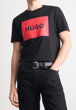 Homme portant un T-shirt noir avec un grand carré rouge et le texte "HUGO", associé à un pantalon noir, une ceinture et une bague argentée à la main gauche.