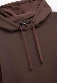 Brązowa bluza z kapturem o teksturowanej tkaninie, z kieszenią z przodu i akcentami w postaci sznurków. Metka widoczna wewnątrz dekoltu.