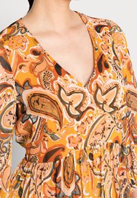 Kobieta w sukience z dekoltem w serek, w pomarańczowe, różowe, brązowe i zielone wzory kwiatowe i paisley, z detalem w talii w formie zgromadzenia materiału.