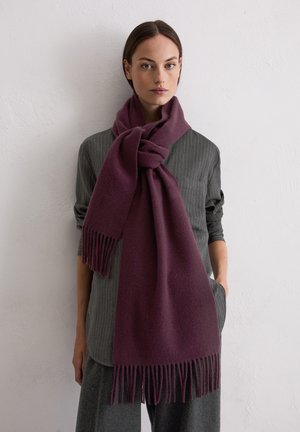 Femme portant une écharpe à franges violet foncé nouée en un nœud lâche sur une chemise grise à fines rayures et un pantalon gris, debout contre un mur blanc.