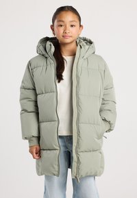 Niña joven con cabello largo y oscuro, vistiendo una chaqueta acolchada con capucha de color verde claro, una camisa blanca y jeans de color azul claro, de pie contra un fondo liso.