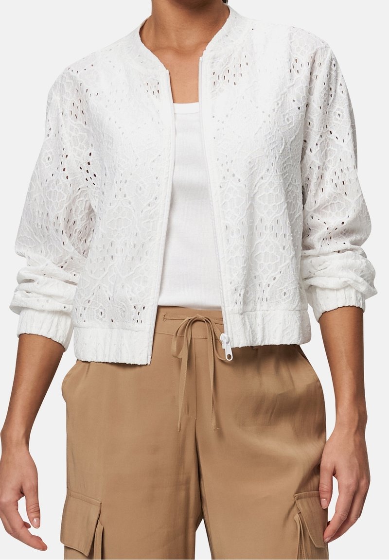 Veste blanche brodée zippée avec poignets élastiques portée sur un haut blanc, associée à un pantalon cargo beige avec taille à cordon.