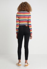 Suéter de rayas acanalado en colores vibrantes, jeans negros ajustados, zapatillas casuales con acentos amarillos. La vista posterior muestra la textura y los detalles del diseño.