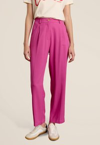 Felroze high-waisted broek met een ontspannen pasvorm, voorzien van voorplooien, twee zijzakken en een knoopsluiting. Draagt met witte sneakers.