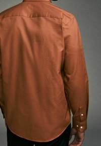Braunes Langarmhemd aus glattem Stoff, mit rundem Saum, geknöpften Manschetten und feiner Struktur. Rückansicht gezeigt.