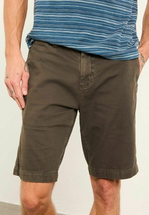 Muž v hnedomi krátkych šortkách do kolien a modrej pruhovanej košeli, stojaci s rukami v vreckách na jednoduchom pozadí.