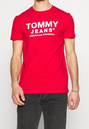 Mand iført en rød T-shirt med korte ærmer med teksten "TOMMY JEANS AMERICAN ORIGINAL" i hvid, sammen med sorte bukser og et sølvarmbånd.