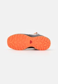 Zapato de exterior con una parte superior texturizada negra y suela de goma naranja brillante que presenta patrones de banda de rodadura profundos para tracción. Incluye detalles reflectantes.