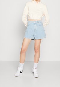 Ljusrosa kort zip-tröja med vit logotyp, ihop med högmidjade blå denimshorts. Vita och svarta högsneakers fulländar looken.