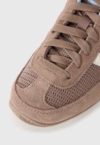 Brūni zamšādas sneakers ar sieta detaļām, teksturētu virsmu, tradicionālu šņorēšanas dizainu un gumijas zoli. Akcentēti ar gaišzilām detaļām.