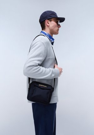 Man in grijze trui, blauwe pet en zonnebril draagt zwarte crossbodytas, staand in profiel tegen een simpele lichte achtergrond.