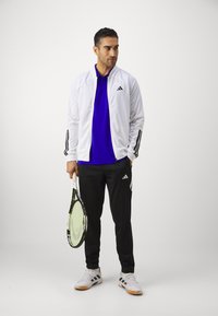 Witte sportjack met zwarte strepen, paarse poloshirt en zwarte broek. Houdt een tennisracket vast en draagt witte schoenen met zwarte accenten.