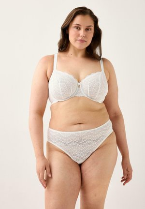 Nainen, jolla on valkoinen pitsirintaliivit ja matching alushousut, seisoo vaaleaa taustaa vasten, kasvot eteenpäin rentoutuneella ilmeellä.