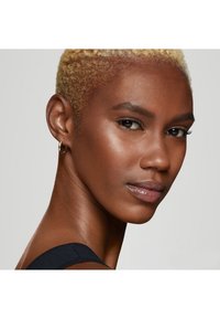 Profilbilde av en person med kort blond hår, brun hud og en liten hoop-ørering. Glatt hud med en subtil glans, iført en mørk topp.