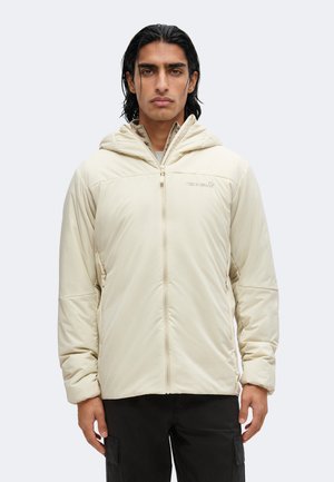 Joven con cabello negro liso viste chaqueta beige con cremallera y capucha y pantalones negros, de pie sobre fondo gris claro.