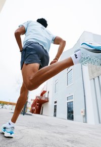Sportsløbesko i lyseblå og hvid, parret med grå shorts og en lyseblå skjorte, båret af en person i bevægelse.