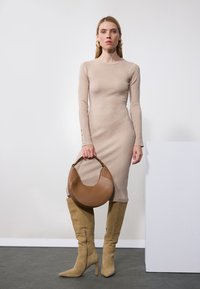 Beige ribbestrikket kjole med lange ermer og knappdetaljer, sammen med knehøye beige semskede støvler og en rund brun håndveske.