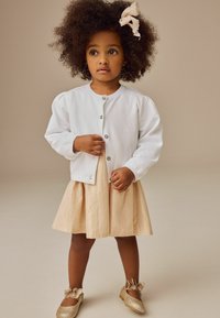 Cardigan en coton blanc avec des manches bouffantes, jupe plissée beige et chaussures dorées pailletées avec nœuds. L'enfant se tient devant un fond neutre.