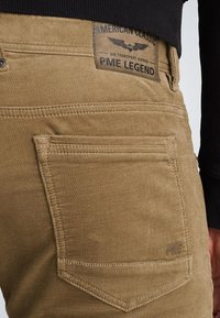 Pantalones de algodón color topo con un acabado texturizado, que cuentan con un bolsillo trasero y una etiqueta con "PME Legend". Se aprecian detalles cosidos a lo largo de las costuras.