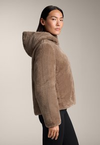 Brun imitationspäls jacka med huva, boxig silhuett, långa ärmar och mjuk textur. Matchas med svarta leggings för en komplett look.