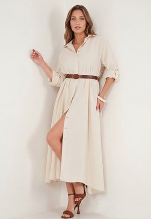 Donna che indossa un vestito camicia beige abbottonato con cintura marrone e sandali con tacco alto e cinturini intrecciati, posando contro un muro bianco.