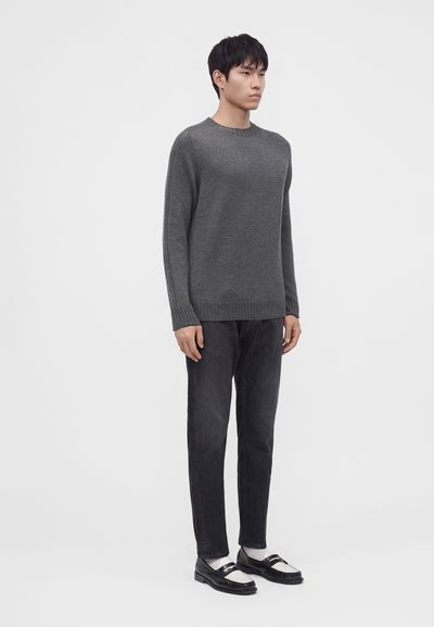 Jeune homme portant un pull gris foncé, un jean noir, des chaussettes blanches et des mocassins noir et blanc, debout devant un fond uni clair.