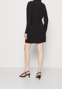 Robe noire à manches longues avec une coupe ajustée, une texture lisse et une discrète couture au dos. Associée à des sandales à talons hauts noires avec brides.