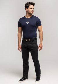 Mężczyzna w granatowej koszulce z logo, czarnych dżinsach, czarnym pasku i czarnych trampkach, stojący i patrzący w bok na jednolitym tle.