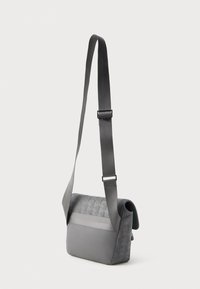 Bolsa de ombro cinza com alça ajustável e padrão geométrico em relevo no flap e nos lados, apresentada contra um fundo liso.