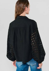 Eine Frau mit langen braunen Haaren trägt eine schwarze Bluse mit Augenlöchern und Puffärmeln sowie blaue Jeans. Sie wird von hinten vor einem einfarbigen Hintergrund gezeigt.
