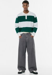 Grün-weißer gestreifter Pullover mit Kragen, graue weit geschnittene Jeans und schwarz-weiße Schuhe. Strukturierter Stoff mit sichtbaren Nähten und Taschen.