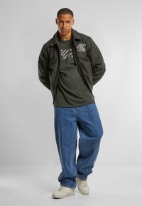 Dark green fleece jacket over a green t-shirt, paired with loose, light blue denim jeans and white sneakers. Simple, casual design.